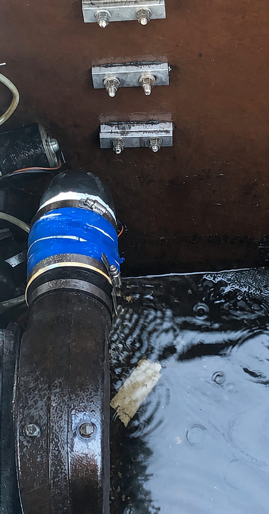 The finished repair: foil and a flattened can sleeved over the exhaust hose, wrapped in blue electrical tape and held by two stainless hose clamps, with standing water in the bilge below
