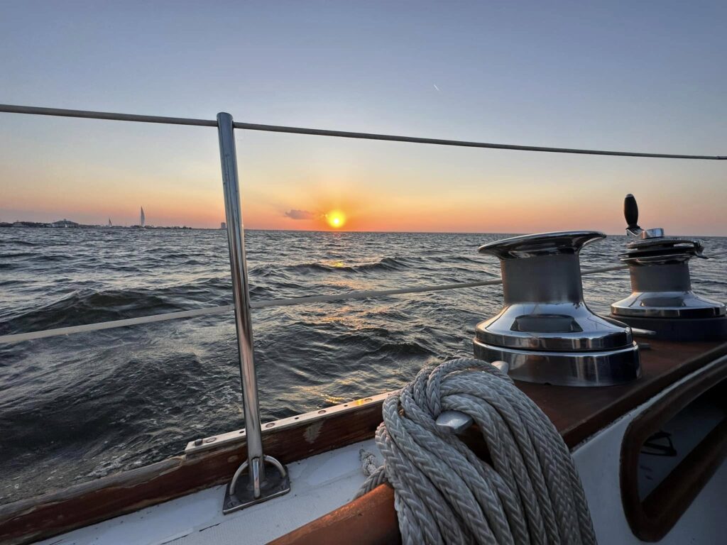 Sunset over Lake Pontchartrain seen from the cockpit of a sailboat, past a teak rail, two winches and a coiled sheet, with another boat's rig on the horizon