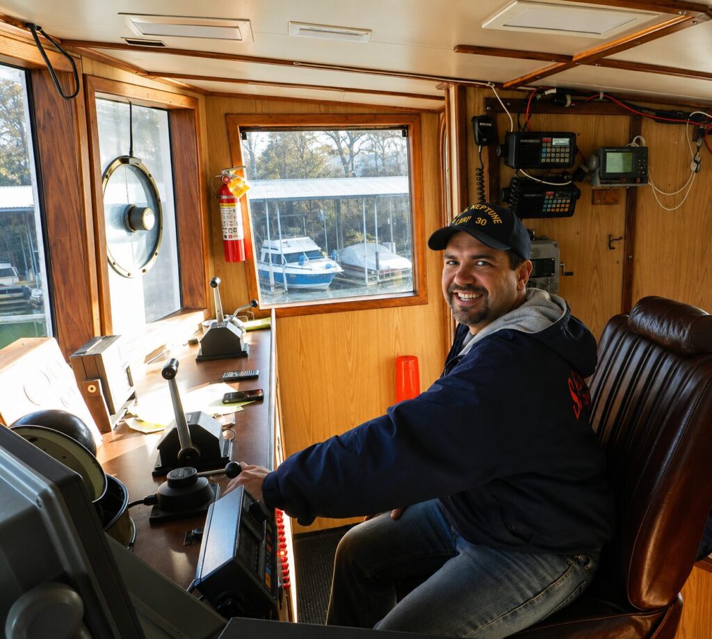 Nate Eckler at the helm of the 95-foot fishing vessel Emerald Sea, in a varnished wood wheelhouse with two radios, a sounder and the engine controls