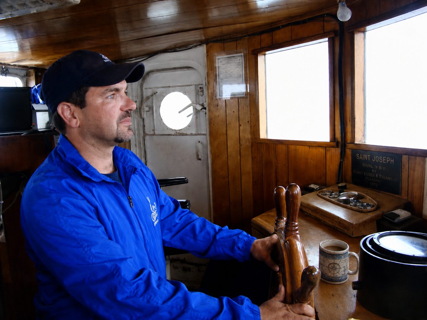 Nate Eckler at the wheel in the wooden wheelhouse of the Saint Joseph, her builder's plaque on the bulkhead behind him