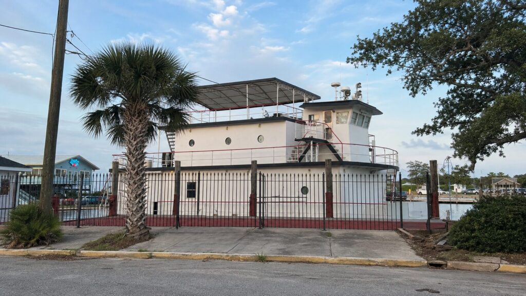 A towboat moored hard against a street behind an iron fence, her wheelhouse and radar masts rising over a palm tree in a residential neighborhood
